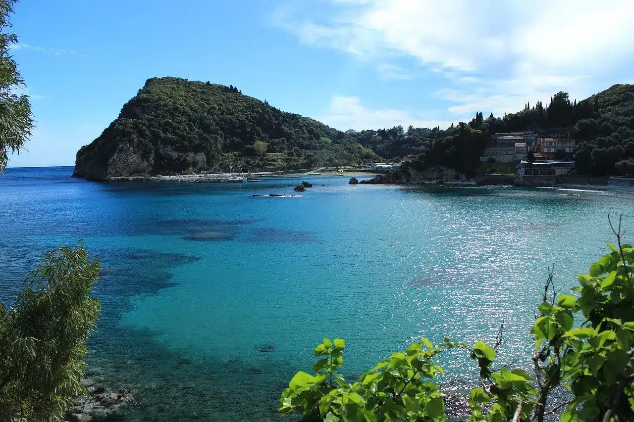 Eine ruhige Meereslandschaft mit einem ruhigen blaugrünen Ozean neben einer üppig grünen Halbinsel unter einem strahlend blauen Himmel. Küstengebäude und eine Segelyacht liegen zwischen Bäumen auf der rechten Seite.