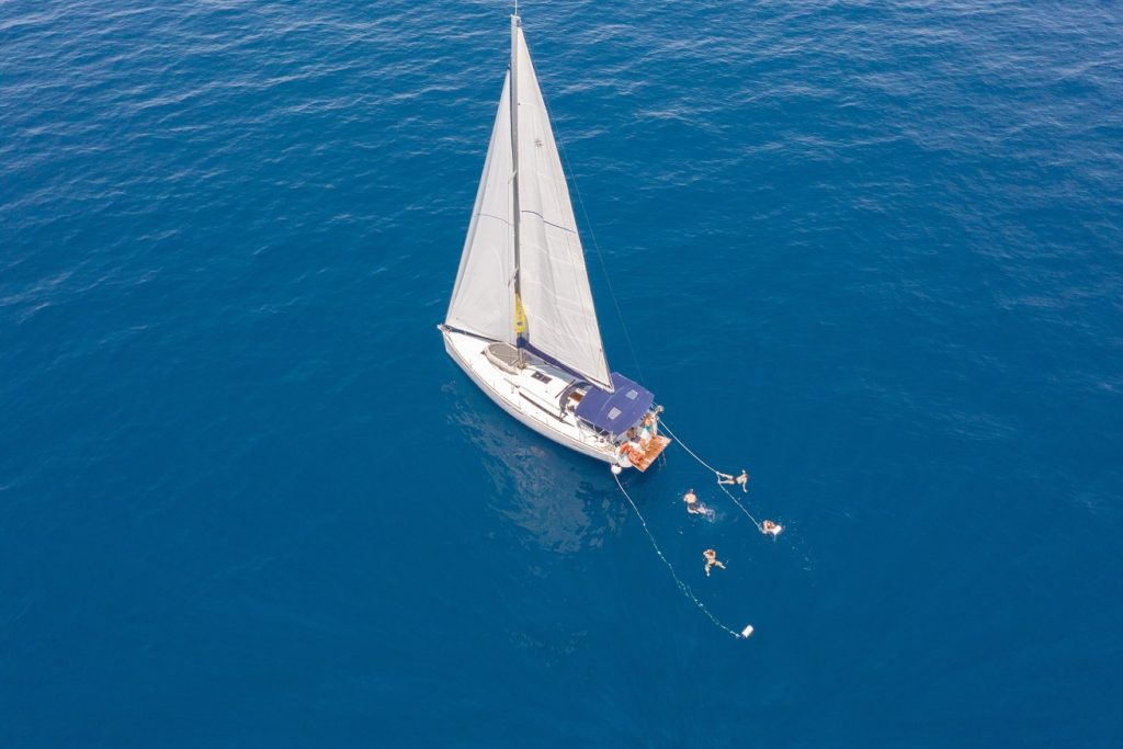 Luftaufnahme eines Segeltörns mit weißen Segeln auf einem klaren blauen Ozean, mit zwei Menschen, die in der Nähe schwimmen.