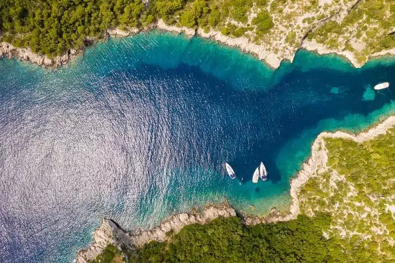 Luftaufnahme einer ruhigen Bucht mit kristallklarem, türkisfarbenem Wasser, umgeben von einer zerklüfteten Küste. Drei Boote sind zu sehen, die während eines Segelurlaubs in Ufernähe treiben.