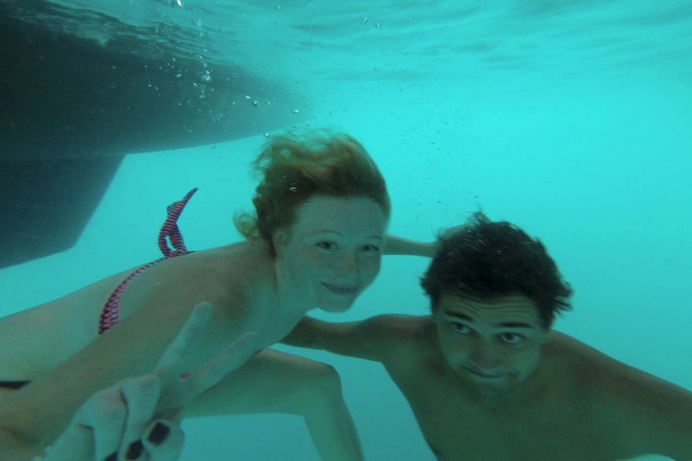 Unterwasserfoto einer jungen Frau und eines Mannes, die in die Kamera lächeln, wobei die Frau mit ihrer Hand ein Peace-Zeichen macht, beide während ihres Segelurlaubs in klares, blaues Wasser getaucht. Wassersportart für Badeurlaub.
