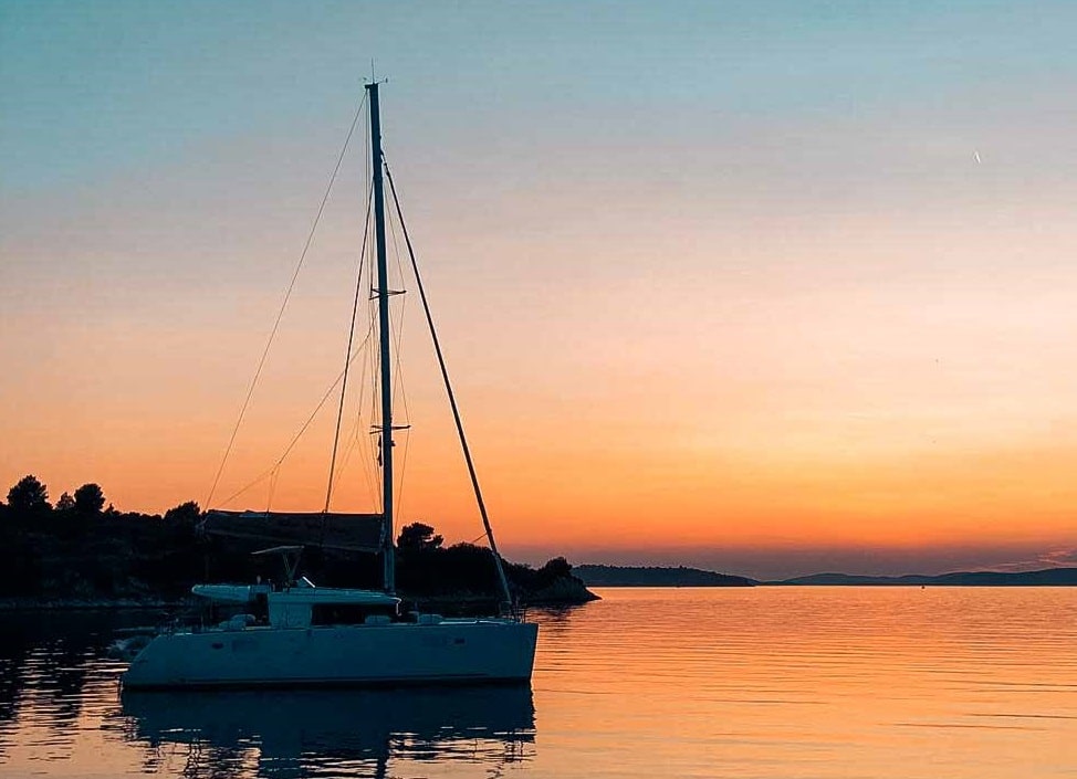 Ein ruhiger Sonnenuntergang mit einem leuchtend orangefarbenen Himmel, der sich im ruhigen Wasser spiegelt und ein Segelboot von Segelreise in der Nähe einer von Bäumen gesäumten Küste hervorhebt. Gute Privatreisen in Top-Ziele.