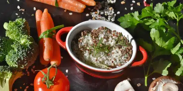 Eine kleine Schüssel mit gewürztem Hackfleisch, umgeben von frischem Gemüse wie Karotten, Tomaten, Brokkoli und Pilzen auf einer dunklen Oberfläche einer Segelyacht.