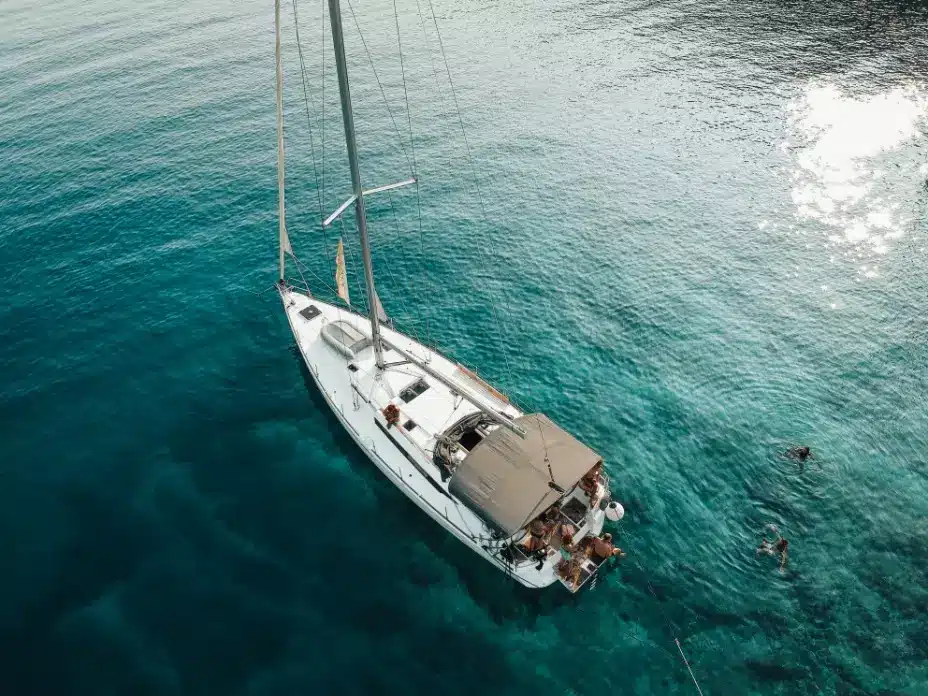 Luftaufnahme einer weißen Segelyacht auf einem klaren, blauen Meer mit Menschen, die auf dem Deck ein Sonnenbad nehmen, und einer Person, die während ihres Segeltörns in der Nähe schwimmt.