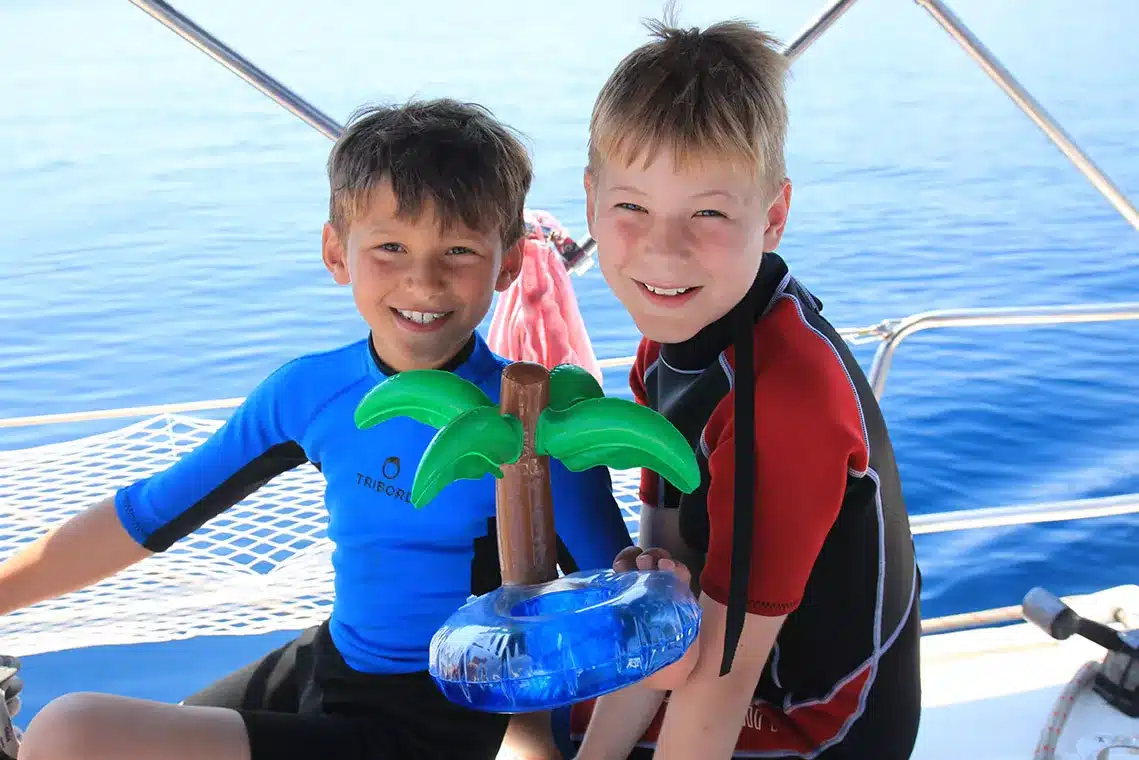 Zwei Jungen mit freudigem Gesichtsausdruck und Schwimmwesten halten eine aufblasbare Palme auf einem Boot, während sie unter klarem blauen Himmel segeln. Familien Badeurlaub Tipps