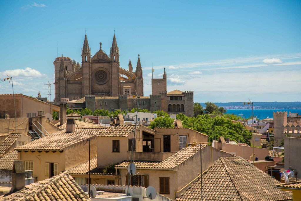 Ein malerischer Blick auf eine historische Kathedrale mit kunstvollen Fassaden, die während eines Segelurlaubs unter einem klaren blauen Himmel die Skyline hinter einer Ansammlung von Häusern mit Terrakotta-Dächern dominiert.