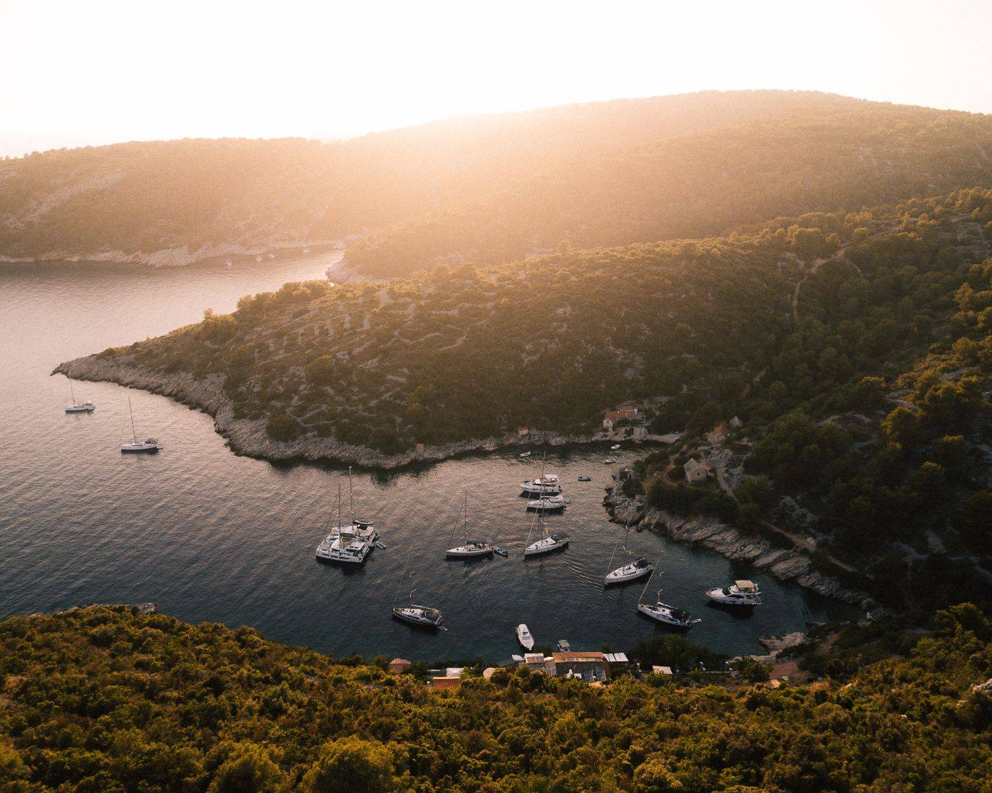 Eine ruhige Bucht bei Sonnenuntergang, mit mehreren Yachten und Booten, die in Küstennähe vor Anker liegen. Das Wasser schimmert, während das goldene Sonnenlicht über das hügelige, mit dichtem Grün bedeckte Gelände strömt. Diese ruhige Umgebung, perfekt für einen Katamaran-Segeltörn, vermittelt ein Gefühl friedlicher Abgeschiedenheit und natürlicher Schönheit.