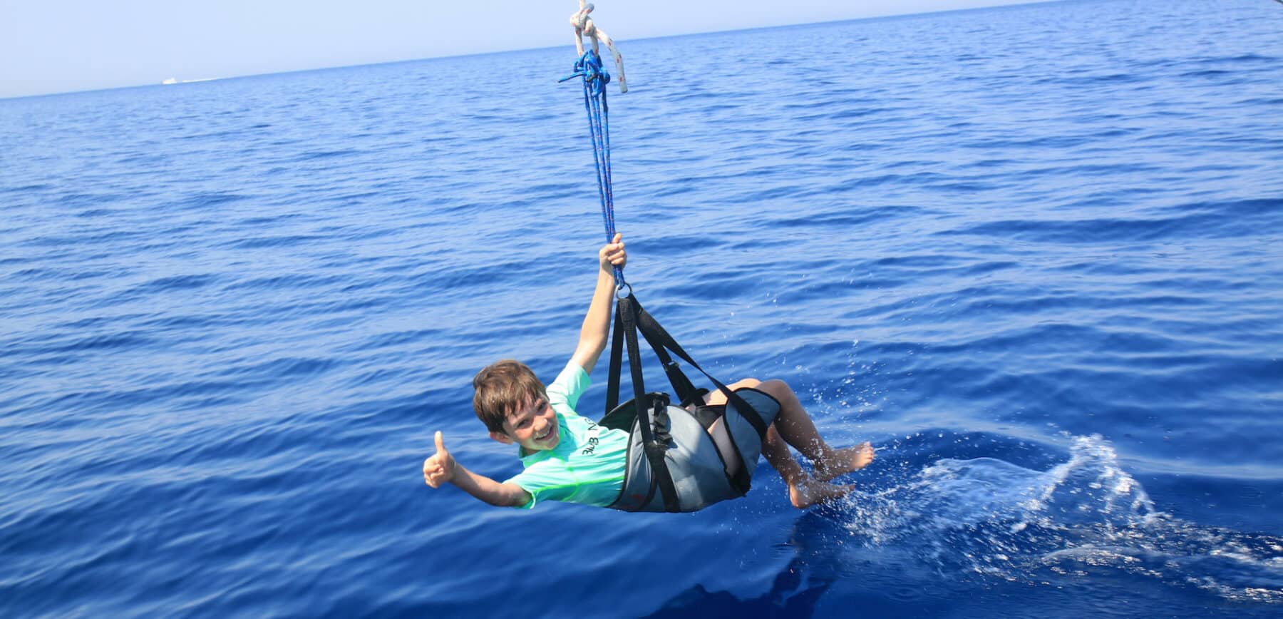 Ein Mann in grünem Hemd und Shorts genießt das Parasailing über dem tiefblauen Meer und streckt dabei seinen Daumen nach oben, während er unter einem an einem Katamaran befestigten Fallschirm hängt.