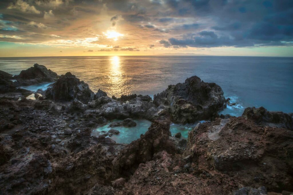 Ein dramatischer Sonnenuntergang über einer zerklüfteten Küste mit Felsformationen und Gezeitentümpeln, in denen sich der Himmel spiegelt, vor der Kulisse von Wolken und ruhiger See, auf der eine Segelyacht anmutig dahinsegelt.