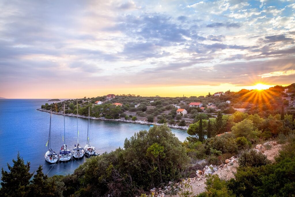 Blick auf den Sonnenuntergang über einem ruhigen Hafen mit mehreren Segelbooten, umgeben von üppigem Grün und einem kleinen Dorf, unter einem weiten, farbenfrohen Himmel während eines Segeltörns.