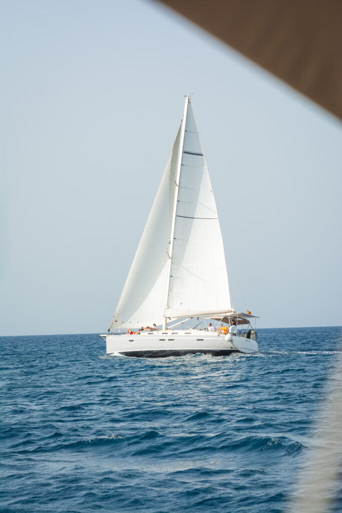 Ein weißes Segelboot mit vollen Segeln gleitet über ein ruhiges blaues Meer, betrachtet durch einen unscharfen Vordergrundrahmen während eines Segeltörns.