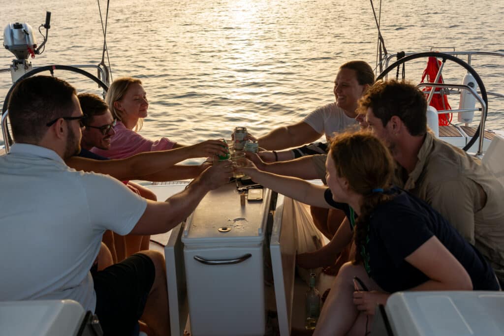 Eine Gruppe von Freunden genießt Getränke und lacht gemeinsam auf einer Segelyacht bei Sonnenuntergang, während sich das goldene Licht im Meer spiegelt.