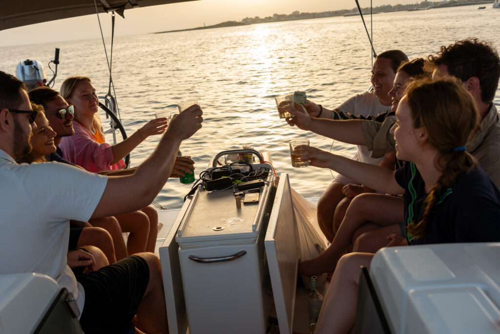 Eine Gruppe von Freunden genießt bei Sonnenuntergang Drinks auf einer Segelyacht und stößt in fröhlicher Atmosphäre gemeinsam an, mit einem malerischen Blick auf das Wasser und den Horizont im Hintergrund.