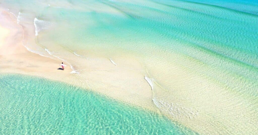 Luftaufnahme einer Person, die an einem unberührten Sandstrand entlangspaziert, an dessen Ufer kristallklares, türkisfarbenes Wasser sanft plätschert. Die Szene fängt die ruhige und weite Schönheit des Strandes ein und weckt Gedanken an eine Segelreise. Traumstrand in der Karibik. Badeurlaub