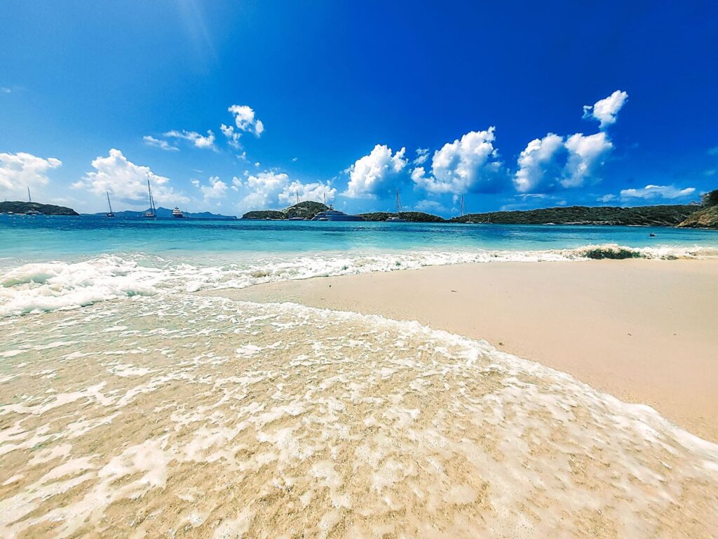 Eine ruhige Strandszene mit sanften Wellen, die unter einem strahlend blauen Himmel an das Sandufer plätschern. Im Hintergrund ankern Katamarane und Segelboote in der Nähe üppiger grüner Hügel. Flauschige weiße Wolken sind über den Himmel verstreut und verstärken die idyllische Küstenatmosphäre. Karibik Segeltörn in der Nebensaison