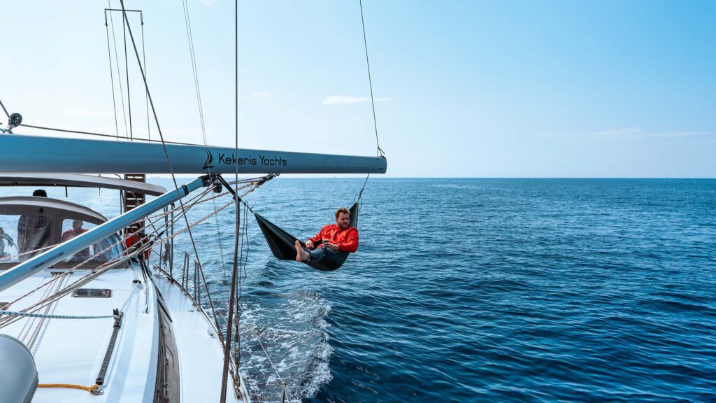 Eine Person in roter Jacke und schwarzen Shorts entspannt sich in einer Hängematte, die am Baum einer Segelyacht mit dem Aufdruck