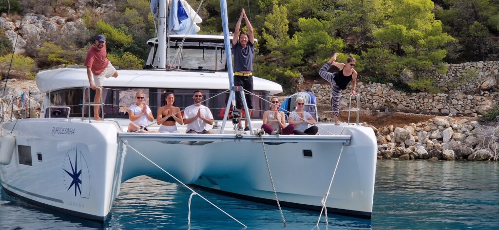Personen die auf einem Katamaran Segelboot Yoga-Posen machen. Die Menschen schauen glücklich aus und lachen. Im Hintergrund sieht man die grüne Natur und Umgebung. Das Wasser ist blau.