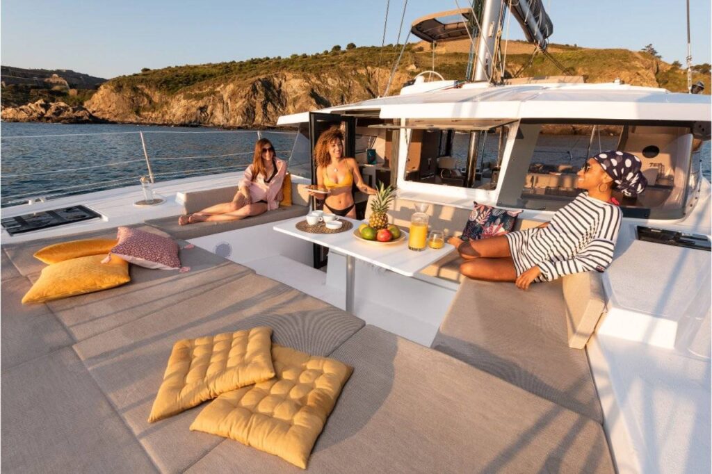Vier Personen entspannen auf einem luxuriösen Youngline-Katamaran mit Bergen und Wasser im Hintergrund. Das Deck ist mit gepolsterten Sitzgelegenheiten und einem Tisch mit Obst und Getränken ausgestattet. Die Atmosphäre ist sonnig und heiter und lässt auf einen gemütlichen Ausflug auf dem Wasser schließen. Die Vorteile von Kleingruppenreisen gegenüber großen Touren