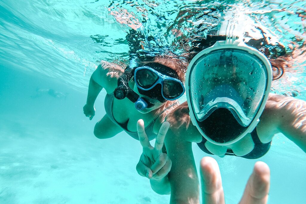 Zwei Personen unter Wasser mit Schnorchelmasken machen während ihres Segelurlaubs ein Selfie. Eine Person hält mit den Fingern ein Peace-Zeichen hoch. Sie sind umgeben von klarem blauem Wasser in der Nähe ihres Katamarans. Abenteuerurlaub