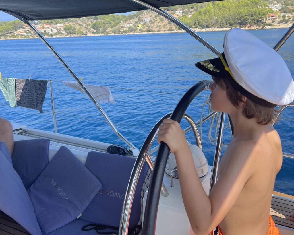 Ein kleiner Junge mit Kapitänsmütze und orangefarbener Badehose steht am Steuer eines Bootes und tut so, als würde er steuern. Das Boot segelt auf klarem, blauem Wasser mit einer hügeligen Küste im Hintergrund und verkörpert den Geist des familienfreundlichen Segeltörns im Mittelmeer. Handtücher und Kleidung trocknen an Bord auf einer Wäscheleine.