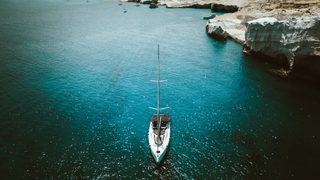 Luftaufnahme eines weißen Segelboots auf ruhigem türkisfarbenem Wasser in der Nähe felsiger Klippen. Das Boot liegt nahe der Küste vor Anker, was an eine ruhige Segeltour erinnert. In der Ferne sind andere kleine Boote zu sehen. Die Szene ist friedlich und sonnenbeschienen, was auf einen angenehmen Tag auf dem Wasser schließen lässt.