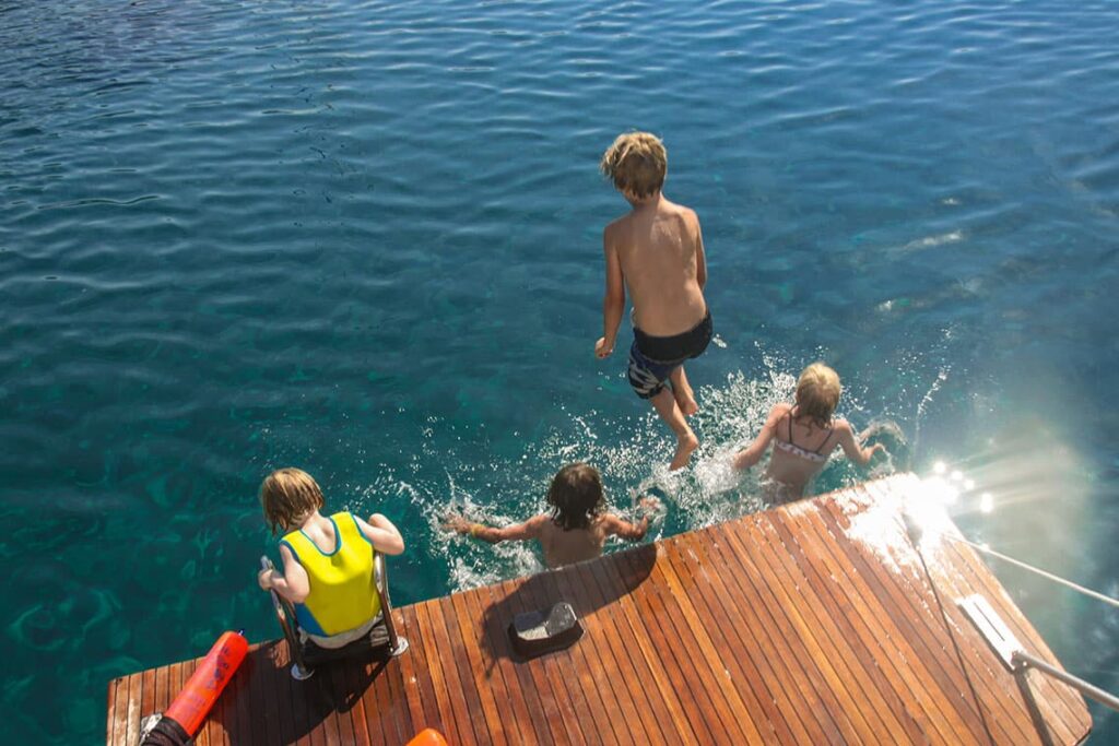 Vier Kinder sind zu sehen, die das Wasser genießen: eines klettert auf eine hölzerne Stegleiter, während die anderen drei gerade springen oder an einem sonnigen Tag im klaren blauen Wasser planschen. Die Szene erinnert an familienfreundliche Segelausflüge im Mittelmeer und steht für Freude und Abenteuer.
