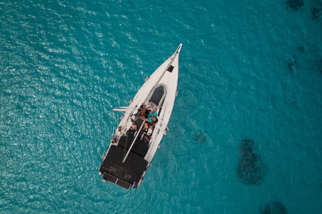 Luftaufnahme eines Segelboots mit einigen Personen an Deck, die während ihrer Segelreise Griechenland in klarem türkisfarbenem Wasser treiben. Der weiße Rumpf des Bootes kontrastiert mit dem leuchtend blauen Meer und schafft eine ruhige und malerische Szene. Mitsegeln in Kleingruppen: Gemeinsam die Meere erkunden