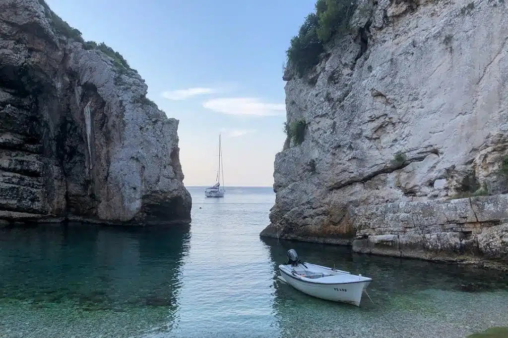 Eine ruhige Bucht mit kristallklarem, türkisfarbenem Wasser, eingerahmt von hoch aufragenden Kalksteinklippen. Ein kleines Boot liegt in Ufernähe und eine Segelyacht ist weiter draußen vertäut.