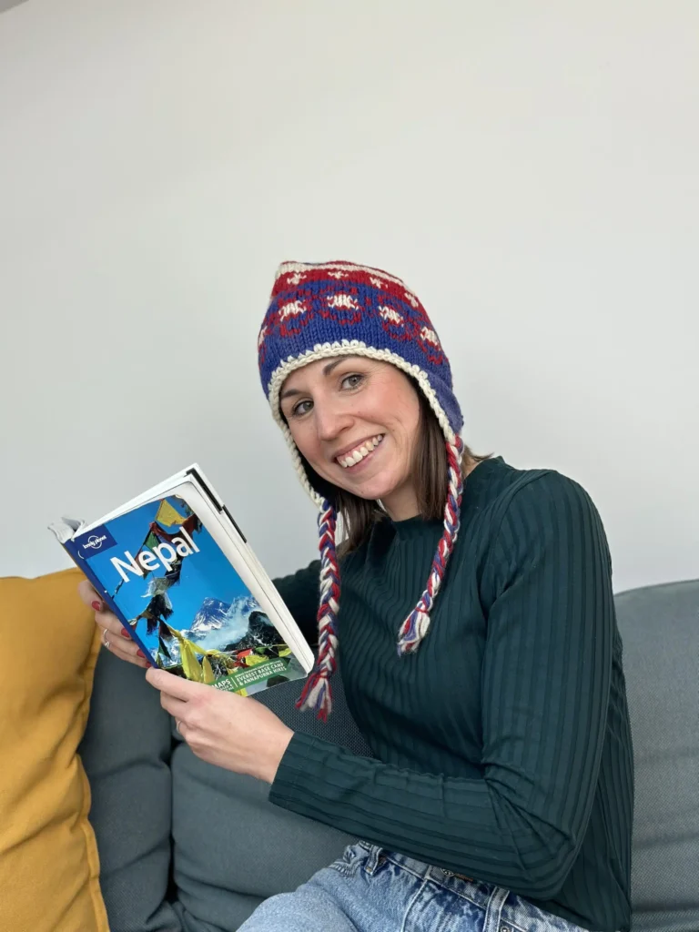 Eine Frau mit einer bunten Strickmütze mit Ohrenklappen und einem grünen Pullover sitzt lächelnd auf einem grauen Sofa. Sie hält einen Reiseführer mit dem Titel 
