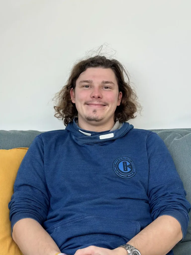 Eine Person mit schulterlangem, welligem Haar sitzt lächelnd auf einem Sofa. Sie trägt ein blaues Sweatshirt mit einem runden Logo darauf. Im Hintergrund sieht man eine hellgraue Wand und ein gelbes Kissen auf der linken Seite des Sofas, was Erinnerungen an den letzten Segelurlaub weckt.
