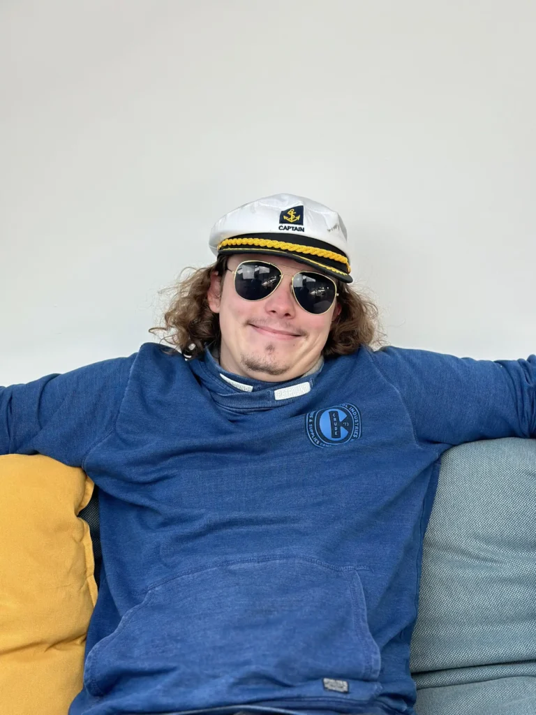 Eine Person mit langen Haaren, einer weißen Kapitänsmütze, Sonnenbrille und einem blauen Sweatshirt mit einem Logo sitzt lächelnd auf einem Sofa und legt die Arme auf die Rückenlehne. Das Sofa hat ein blau-gelbes Kissen. Der Hintergrund ist schlicht weiß und erinnert an die heitere Atmosphäre eines Segeltörn-Abenteuers.