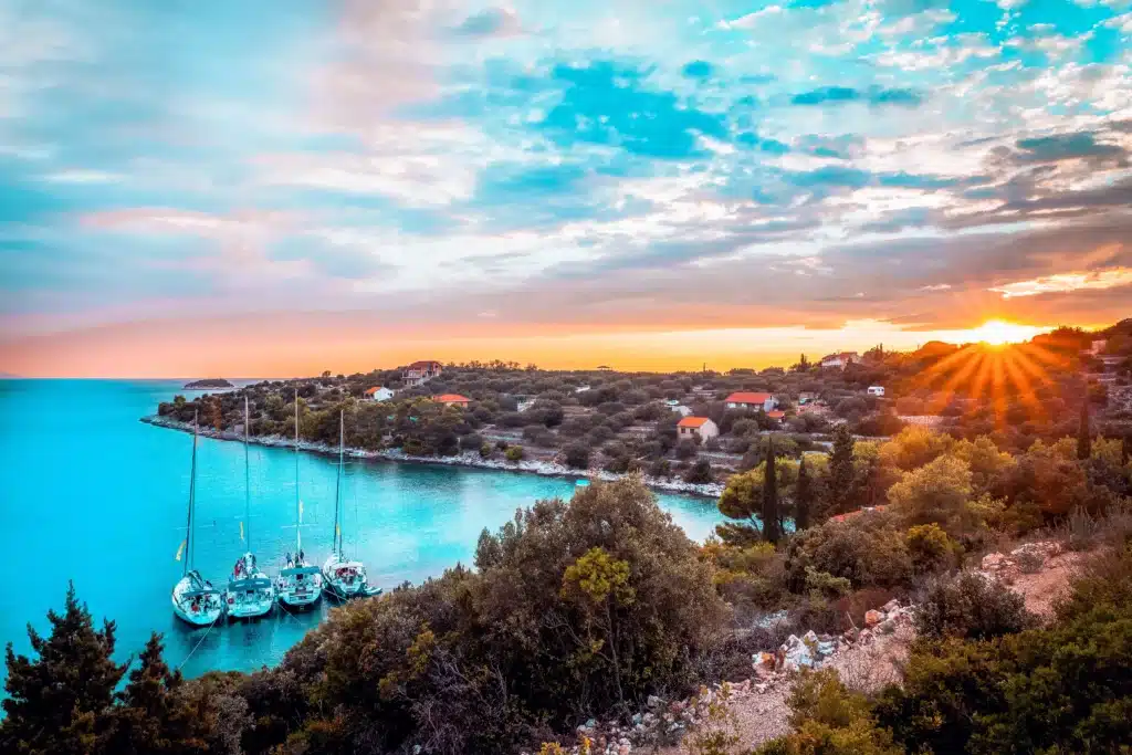 Sonnenuntergang über einer ruhigen Bucht mit Segelbooten, die an einer baumbestandenen Küste festgemacht sind, leuchtende Farben am Himmel, die sich im ruhigen Wasser spiegeln, und ein malerisches Dorf im Hintergrund während eines Segelre