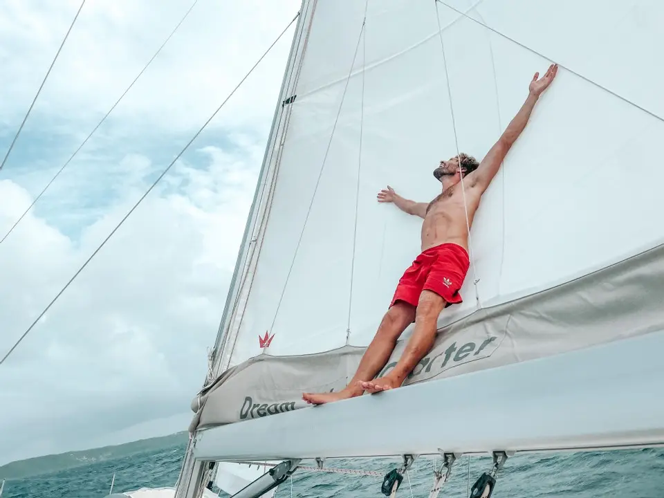 Ein Mann in roten Shorts streckt freudig seine Arme aus, während er auf dem schrägen Segel eines fahrenden Katamarans liegt, über ihm ist der Himmel bewölkt und im Hintergrund das Meer.