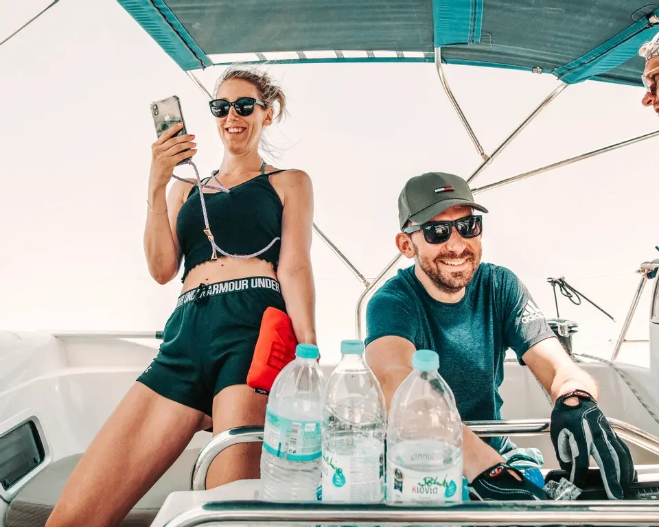 Zwei Menschen genießen einen Segeltörn; eine Frau in Schwarz macht ein Selfie und ein Mann mit Mütze steuert das Boot, umgeben von Wasserflaschen und klarem Himmel.