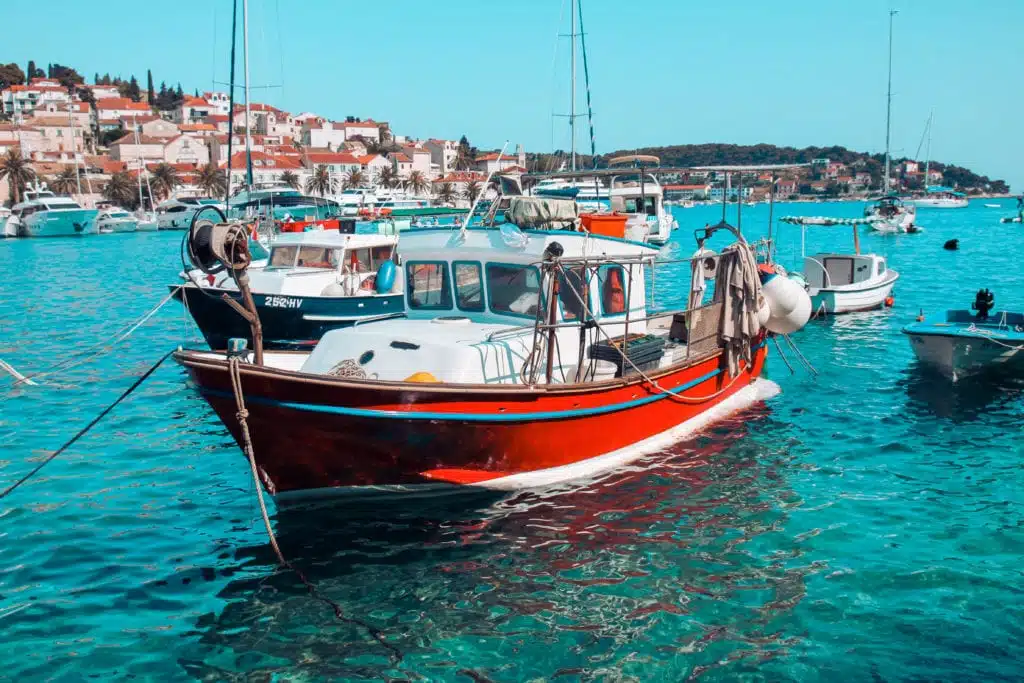 Ein leuchtend rotes Segelboot liegt im klaren, türkisfarbenen Wasser vor Anker. Im Hintergrund sind mehrere andere Boote zu sehen, im Hintergrund ein malerisches Dorf auf einem Hügel unter einem strahlend blauen Himmel.