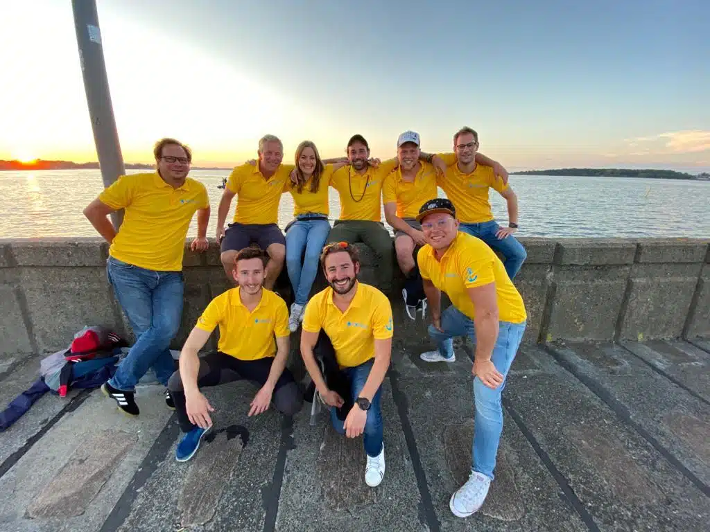 Eine Gruppe von neun Personen in passenden gelben Hemden lächeln und posieren gemeinsam an einem Wasserufer während eines Segeltörns bei Sonnenuntergang.