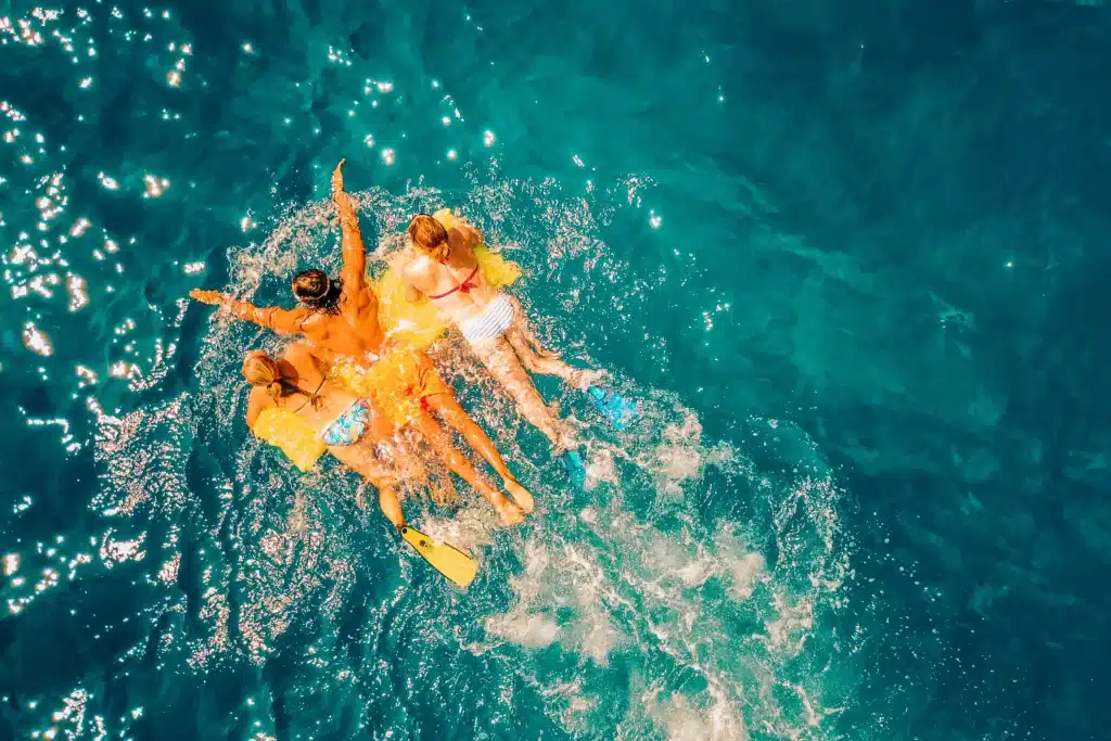 Luftaufnahme von vier Personen, die auf einem Schlauchboot im klaren, blauen Meerwasser treiben, für Spritzer sorgen und einen sonnigen Tag während ihres Segelurlaubs genießen.