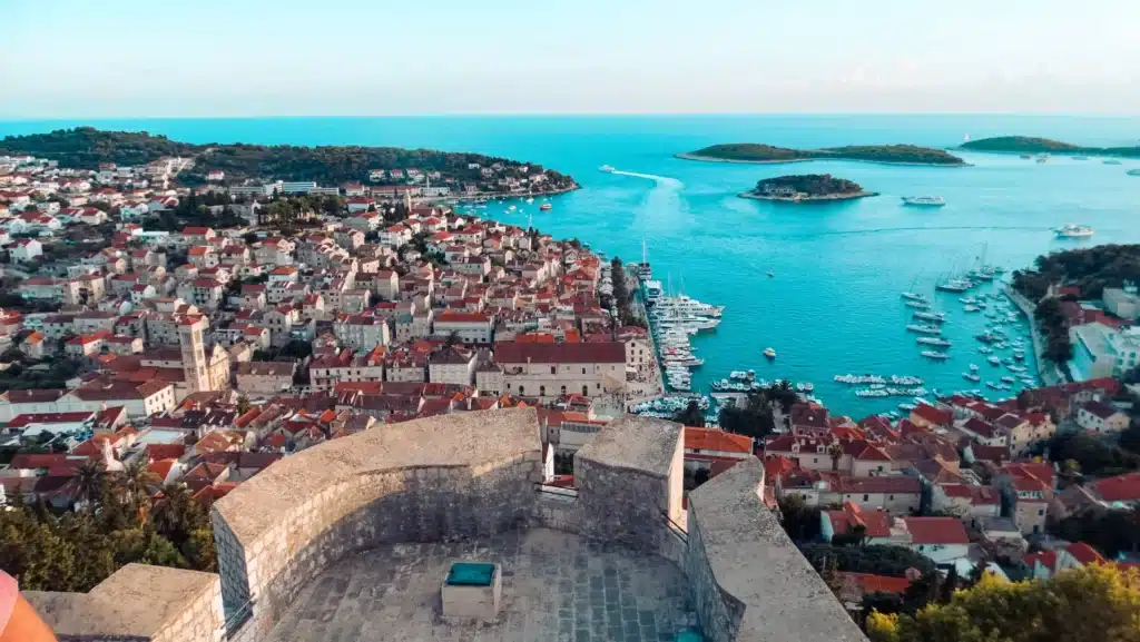 Luftaufnahme einer Küstenstadt mit Terrakotta-Dächern, mit einer historischen Festung im Vordergrund und einem leuchtend blauen Meer mit Segelbooten und Inseln im Hintergrund.