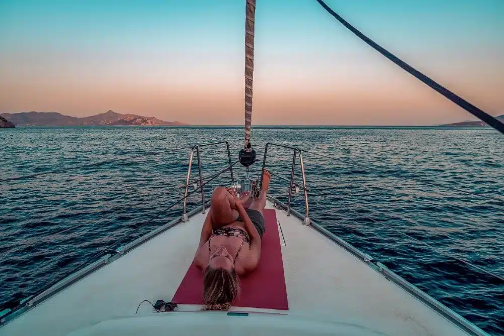 Eine Person entspannt sich während eines Segeltörns bei Sonnenuntergang auf dem Vorderdeck eines Segelboots, mit ruhigem Ozeanwasser und in der Ferne sichtbaren Bergen unter einem farbenfrohen Himmel.