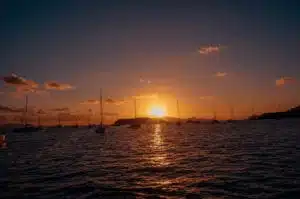 Ein ruhiger Sonnenuntergang über einem Gewässer mit den Silhouetten mehrerer Segelboote, die unter dem leuchtenden Himmel vor Anker liegen und so eine idyllische Segelurlaubsszenerie schaffen.