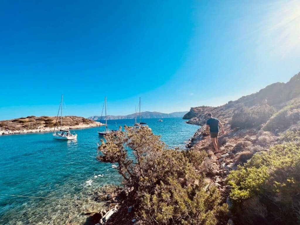 Eine ruhige Küstenszene mit einer Person, die Segelboote betrachtet, die in der Nähe einer felsigen Küste unter einem strahlend blauen Himmel vor Anker liegen. Lebendiges Sonnenlicht erhellt das klare blaue Wasser und die umgebende Landschaft, perfekt für ein Segel