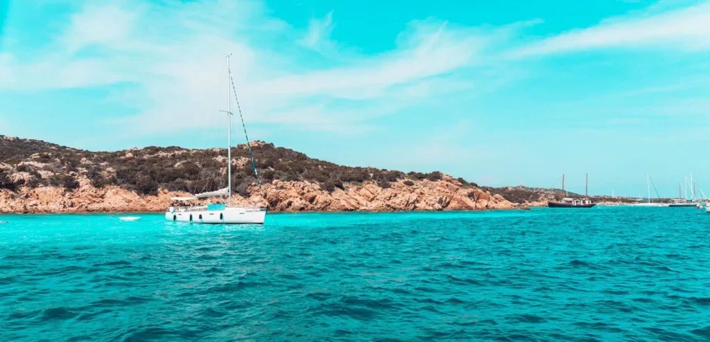 Eine Segelyacht ankert im klaren türkisfarbenen Wasser nahe einer felsigen Küste unter einem strahlend blauen Himmel. In der Ferne sind weitere Segelboote zu sehen.