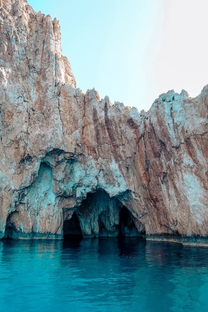 Felsige Klippen am Meer mit einer natürlichen Bogenformation über klarem, türkisfarbenem Wasser. Die Sonne beleuchtet die raue Textur der Felsoberflächen, während eine Segelyacht vorbeisegelt, und verbessert so die malerische Aussicht.