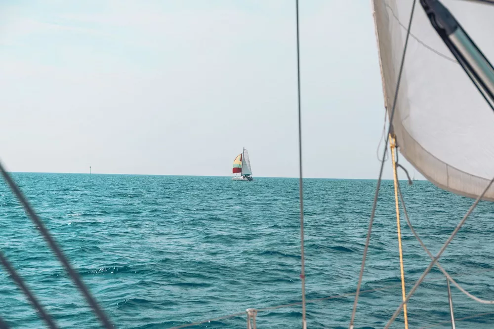 Ein Blick von hinter dem Segel einer Segelyacht auf ein anderes Segelboot mit bunten Segeln am Horizont eines ruhigen blauen Meeres.