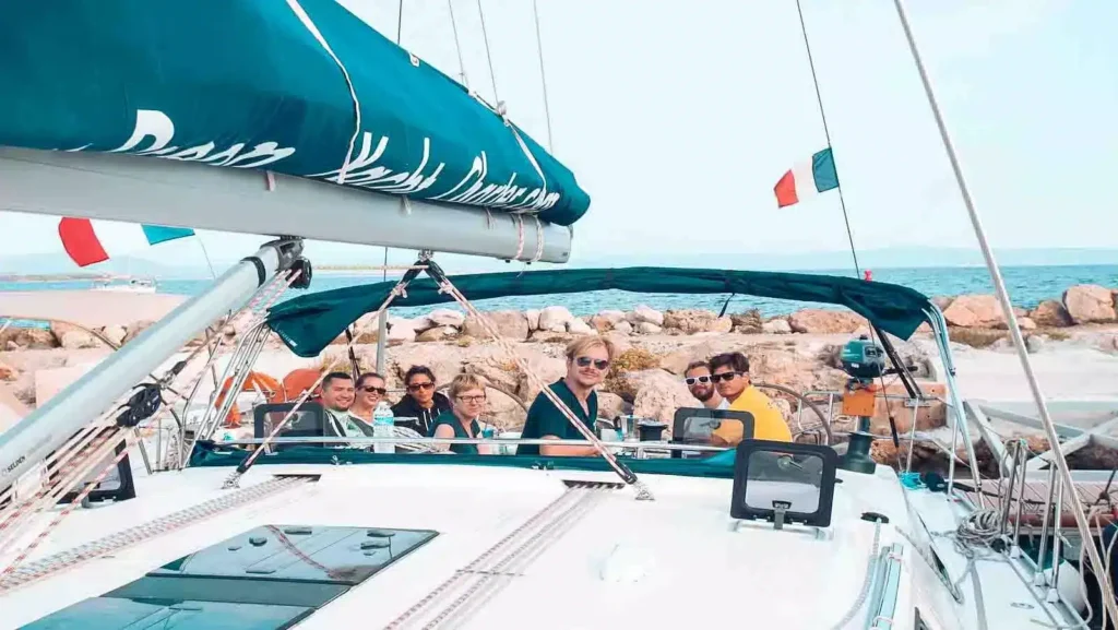 Eine Gruppe von sechs lächelnden Menschen auf einer Segelyacht, nahe einer felsigen Küste unter klarem Himmel. Auf dem Verdeck des Bootes steht 