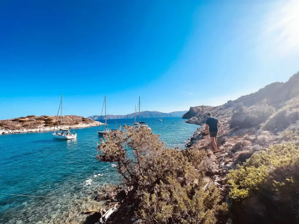 Eine malerische Küstenansicht mit klarem, blauem Wasser und verankerten Segelbooten, darunter ein Katamaran. Felsiges Gelände im Vordergrund, mit einer Person von hinten, die unter einem hellen, sonnigen Himmel auf das Meer blickt.