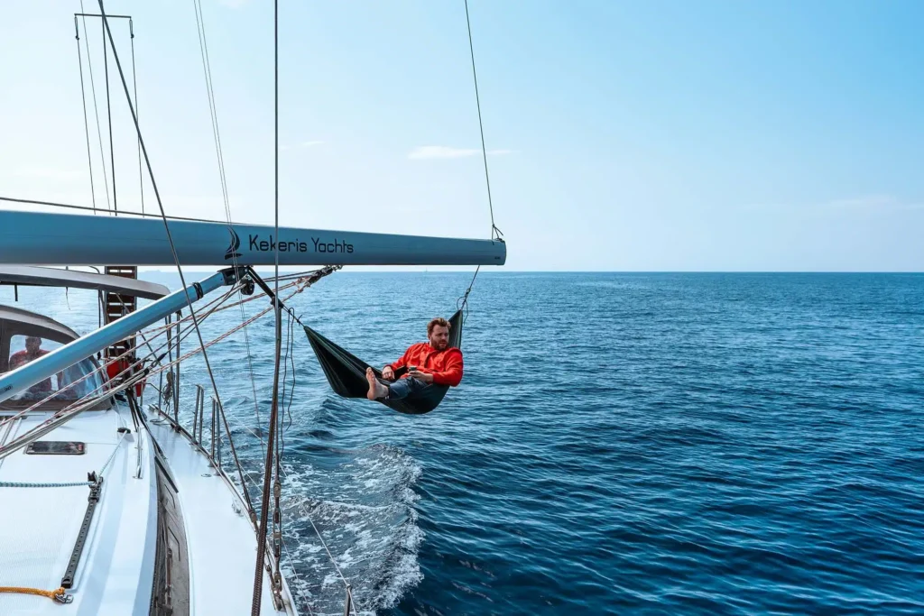 Eine Person in roter Jacke und schwarzen Shorts entspannt sich in einer Hängematte, die am Baum einer Segelyacht mit dem Aufdruck 
