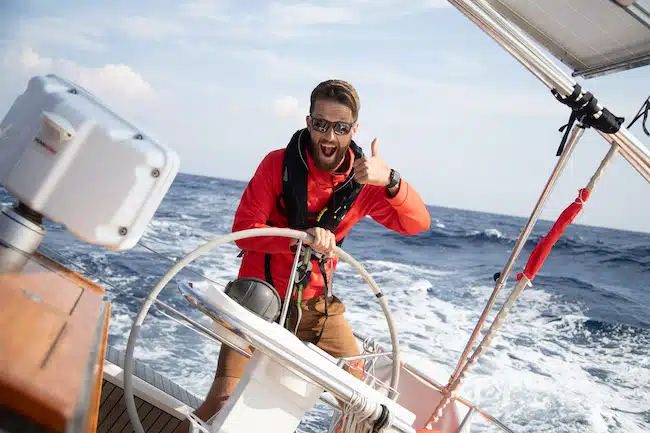 Ein Mann mit Bart, Sonnenbrille und roter Jacke steuert während seiner Segelreise ein Segelboot und streckt den Daumen nach oben, mit dem Meer im Hintergrund.