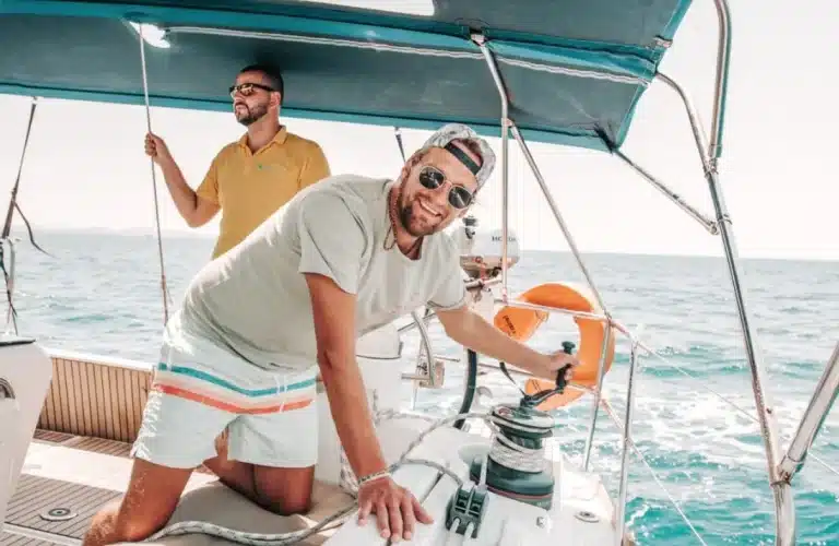 Zwei Männer auf einer Segelyacht; einer steuert das Ruder, der andere steht im Hintergrund. Beide lächeln an einem sonnigen Tag mit Blick auf das Meer.