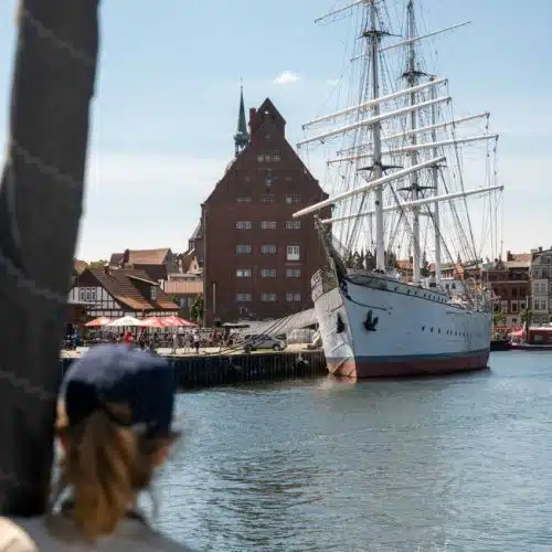 Eine Person mit einer Mütze beobachtet eine große weiße Segelyacht, die an einer sonnigen Uferpromenade vor Anker liegt. Im Hintergrund sind ein historisches rotes Gebäude und andere Strukturen zu sehen.