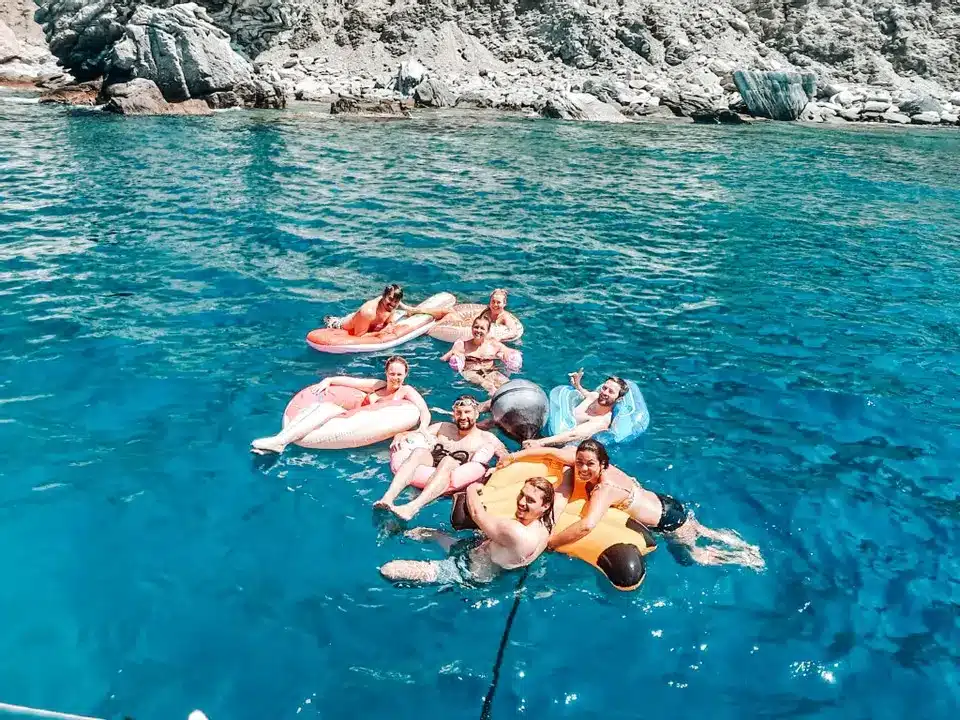 Eine Gruppe von Menschen schwimmt auf einem farbenfrohen Pool im klaren, blauen Ozeanwasser in der Nähe von felsigen Klippen, lächelt und genießt einen sonnigen Tag auf einer Segelyacht.